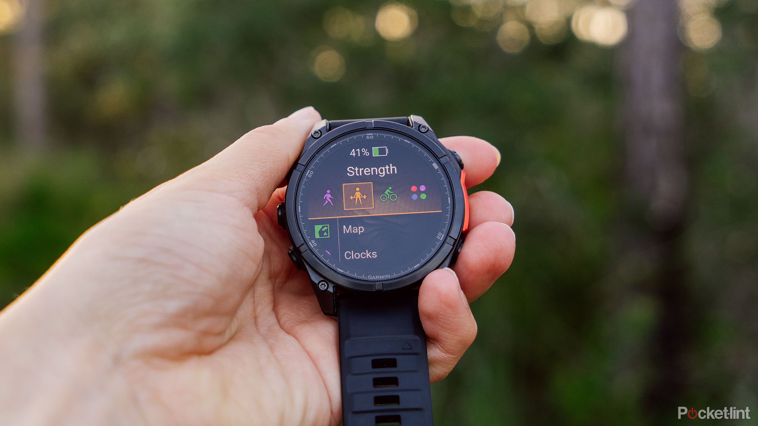 A hand holds the Garmin Fenix 8 watch in front of a blurred forest. 