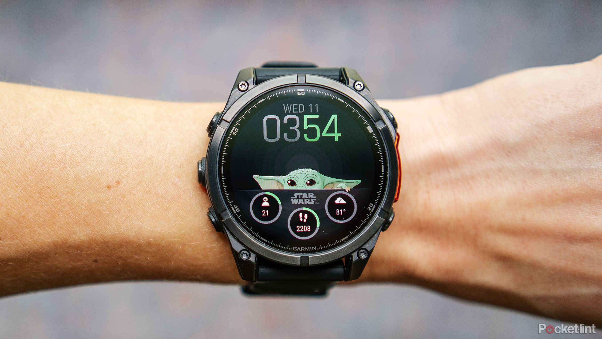 The Garmin Fenix 8 AMOLED with the Grogu watch face is on a wrist held above a blurred blue and red background. 