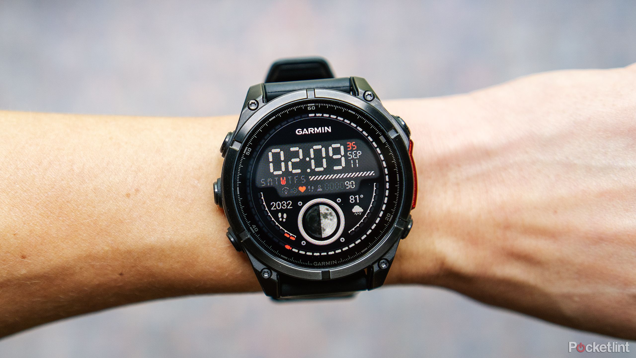 The Garmin Fenix 8 watch with the Garmin Moon Walker watch face is on a wrist held above a blurred blue and red background. 