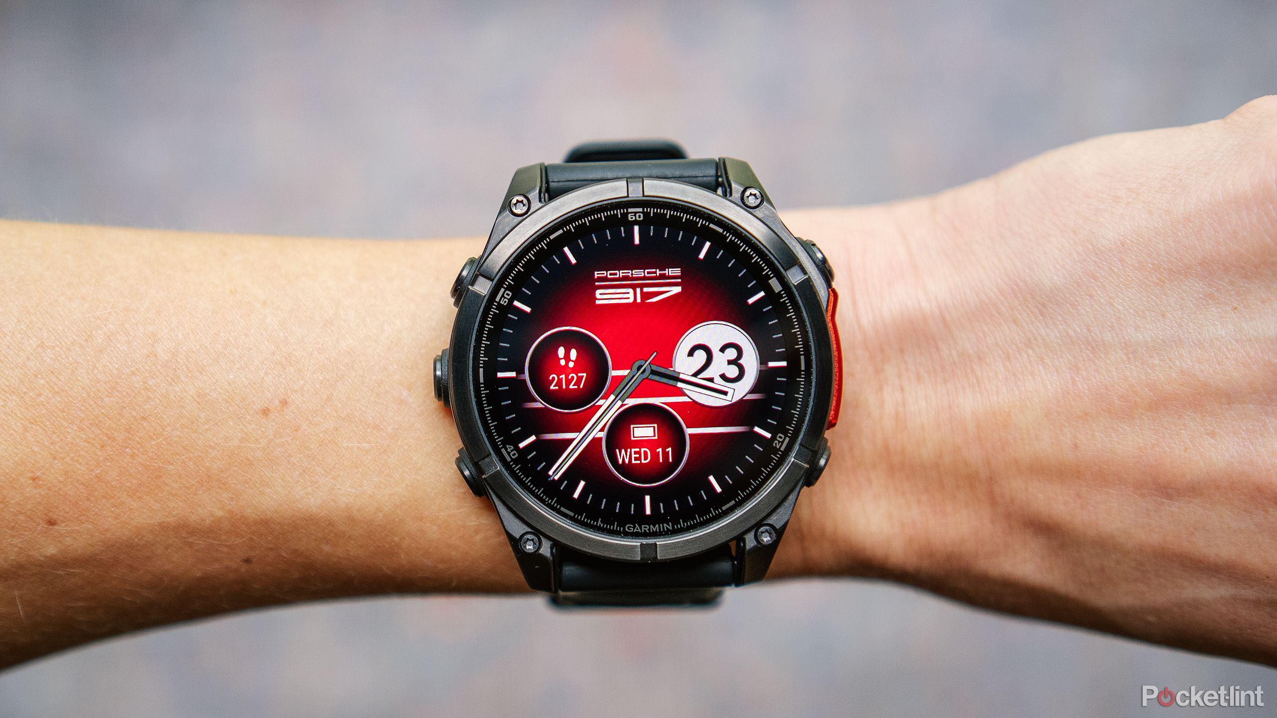 The Garmin Fenix 8 AMOLED with the Porsche watch face is on a wrist held above a blurred blue and red background. 