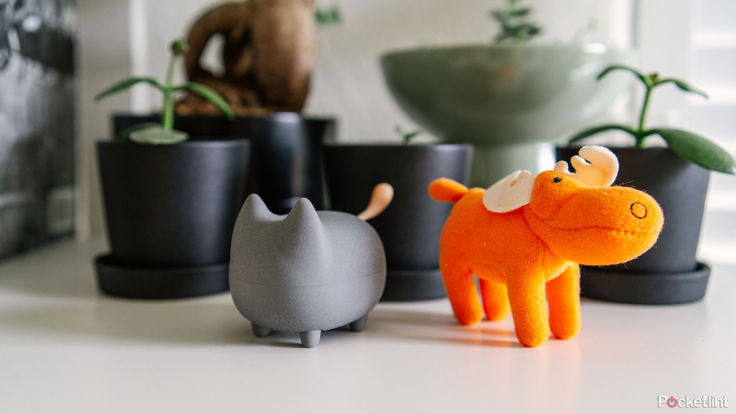 The Gingo Kitty Speaker sits next to an orange stuffed moose on a white shelf in front of potted plants.