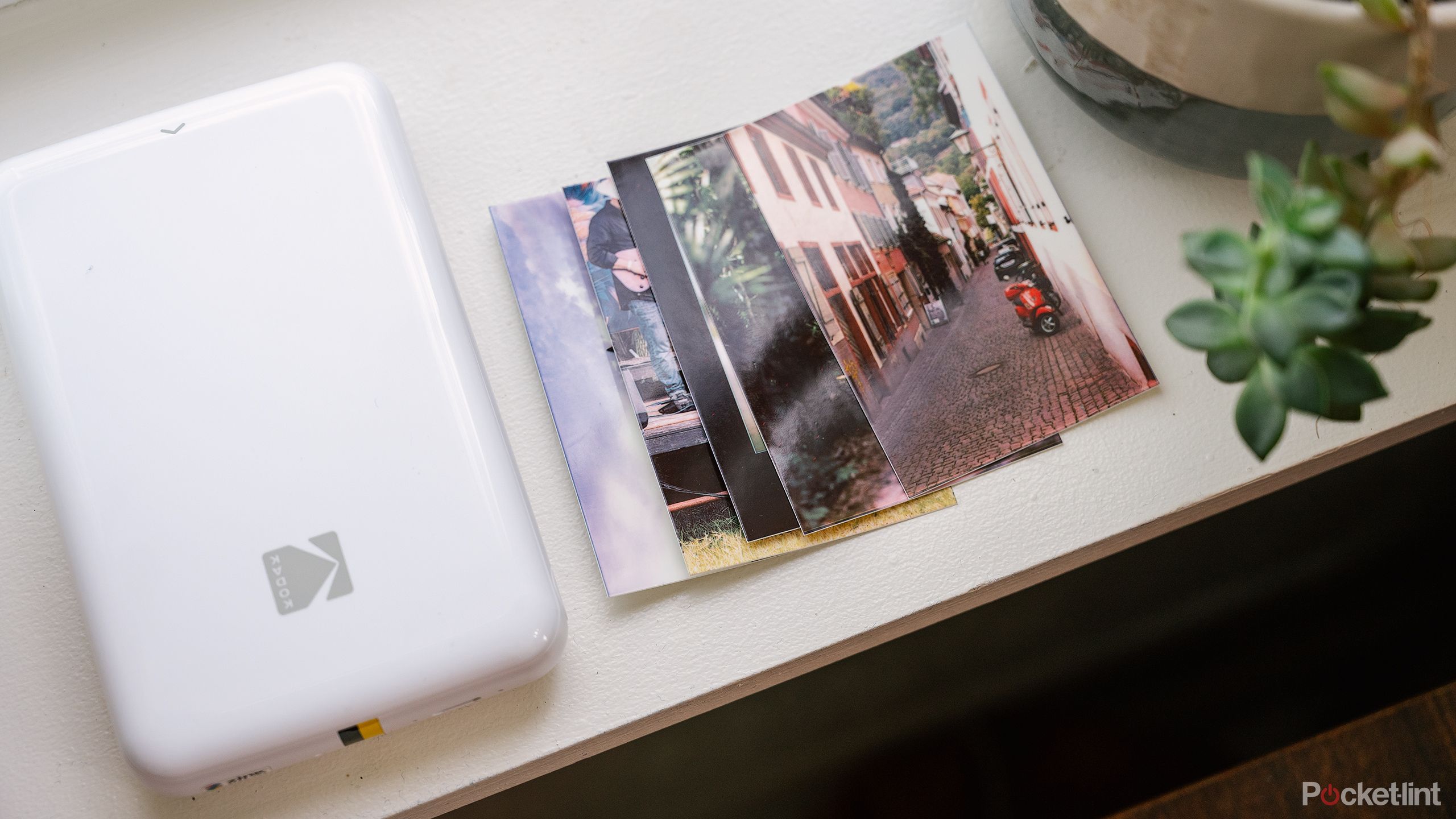 The Kodak Step Mobile Photo Printer and a small pile of prints sits on a windowsill. 