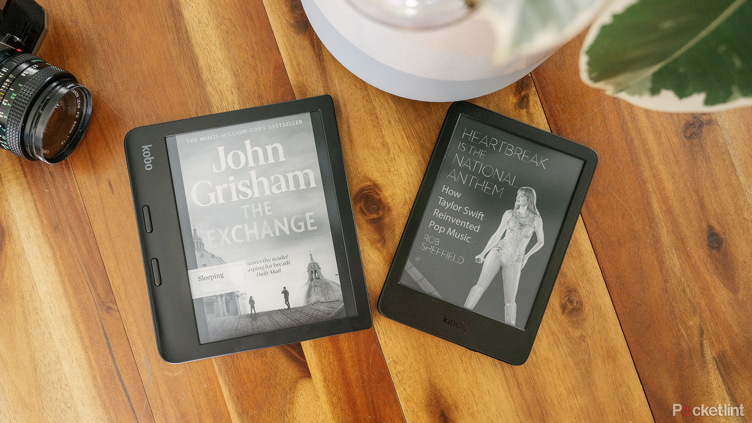 The Kindle Paperwhite sits next to a Kobo Libra 2 on a wood table next to a potted plant and old camera.