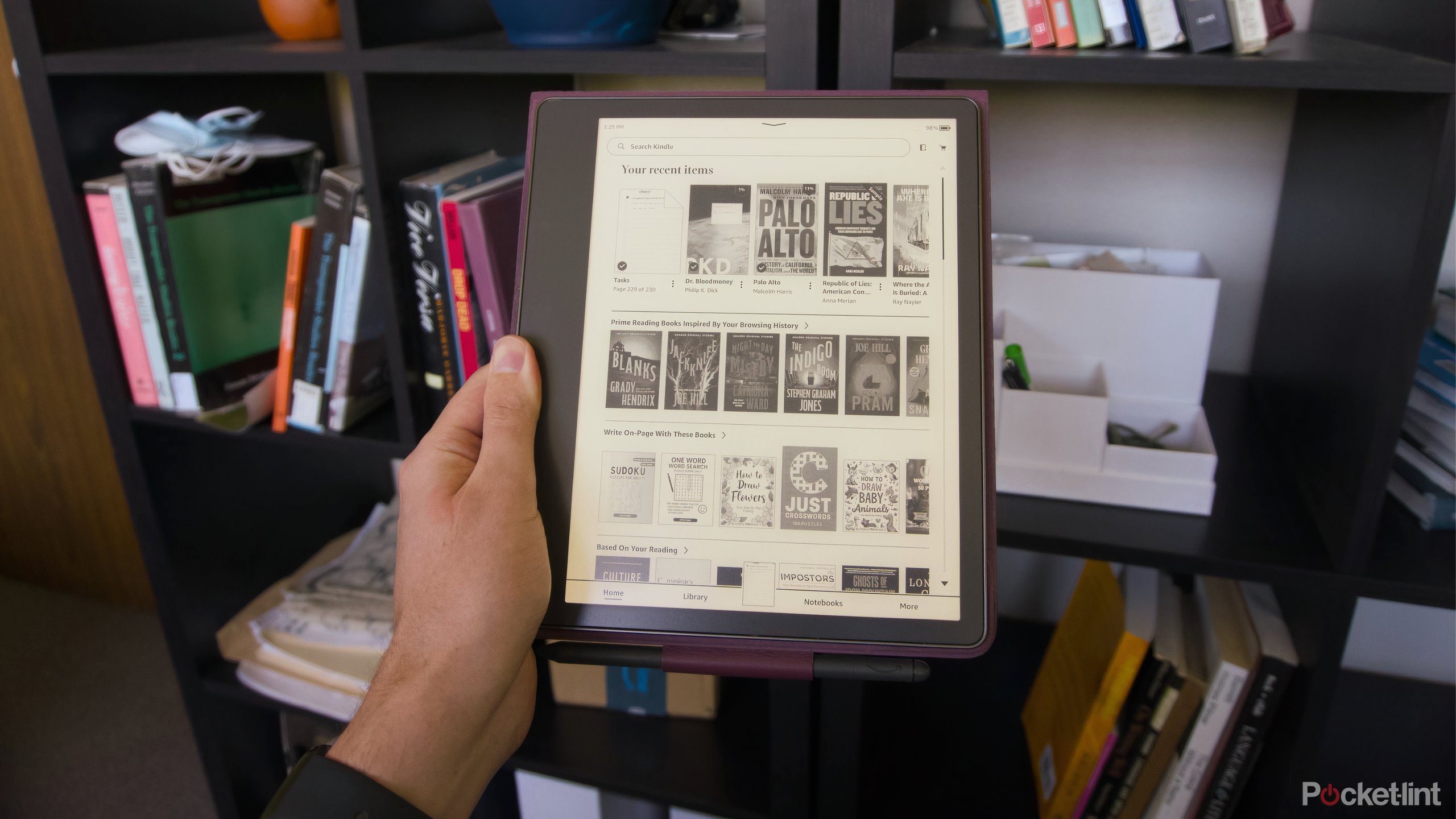 A hand holding a Kindle Scribe in front of a bookcase.
